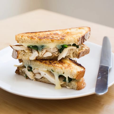 A close-up of a Grilled Chicken Spinach Panini reveals juicy grilled chicken, fresh spinach, and oozing cheese between crusty, buttery bread slices.  